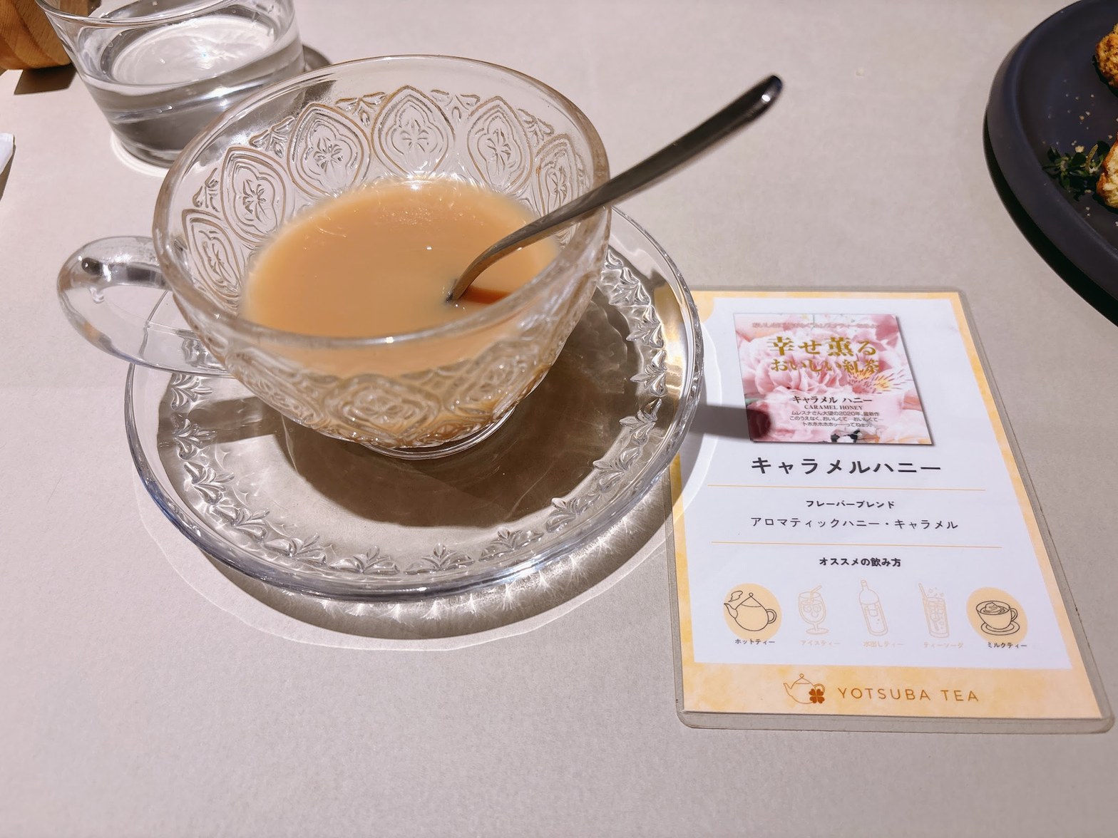カット模様の透明グラスカップに注がれたミルクティー色の紅茶と、隣に立てかけられたYOTSUBA TEAのフレーバーカード「キャラメルハニー」